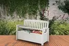 A white storage bench is placed on a wooden deck in a garden setting, surrounded by lush greenery and a stone wall. Náhľad obrázku produktu