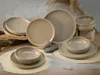 A set of stoneware plates and bowls in a neutral, earthy tone, arranged on a wooden surface with natural elements like dried grass and stones. Produktbild-Vorschau