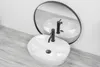 A white oval countertop sink with a black faucet is shown in front of a round mirror reflecting the sink. The sink is placed on a wooden surface. Náhľad obrázku produktu