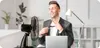 A man in a suit is sitting at a desk in front of a laptop, loosening his tie and fanning himself with his hand in front of a fan. Anteprima dell’immagine prodotto