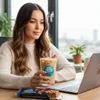 A woman sits at a table with a laptop, holding a glass of iced coffee and a protein bar. Produktbild-Vorschau