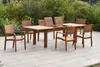 A wooden garden furniture set with a table and chairs is placed on a patio, with lush greenery and some yellow flowers in the background. Produktbild-Vorschau