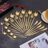 A set of gold-colored cutlery is arranged on a dark gray surface, with two strawberries and a gift box in the background. Produktbild-Vorschau