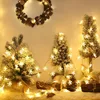 A festive display of Christmas decorations, including small, snow-dusted artificial trees adorned with snowflake-shaped lights, a pinecone wreath, and natural wood elements, all illuminated by warm white lights. Produktbild-Vorschau