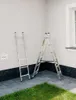 Two ladders are placed on a paved patio next to a white wall. One ladder is leaning against the wall, and the other is in an A-frame position. There is some grass in the foreground and a window on the right side of the wall. Podgląd zdjęcia produktu