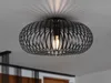 A black, modern ceiling light fixture with a cage-like design is mounted on a white ceiling. The light bulb inside is illuminated, casting a patterned shadow on the ceiling. Náhled obrázku produktu