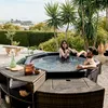A couple relaxing in a hot tub outdoors, with furniture and plants around them. Produktbild-Vorschau