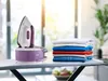 A Braun steam ironing station sits on an ironing board next to a neatly folded stack of clothes. Sunlight streams in from a window in the background. Produktbild-Vorschau