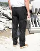 A person wearing black work pants and work boots stands in an outdoor construction or industrial area with rubble and machinery in the background. Anteprima dell’immagine prodotto