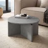 A round coffee table with a stone-like finish is placed on a textured rug in a living room setting, next to a sofa. Voir les images du produit
