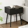 A black bedside table with a drawer and gold handle, placed next to a sofa and a plant. A teapot and cup are on top of the table. Produktbild-Vorschau