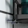 A close-up view of a black metal shower shelf attached to a tiled wall with clear suction cups. Náhled obrázku produktu