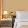 A bedside table with a lamp, flowers, and an alarm clock next to a bed. Produktbild-Vorschau