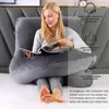A woman is sitting on a bed, reading a magazine, and leaning against a large grey pillow. The pillow is J-shaped and appears to be a pregnancy or nursing pillow. Produktbild-Vorschau