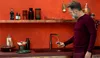 A man is filling a glass with water from a faucet in a kitchen with red walls. Podgląd zdjęcia produktu