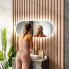 A woman is applying skincare in a modern bathroom with a wooden accent wall and a large illuminated mirror. Produktbild-Vorschau