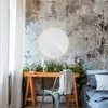A round LED mirror is mounted on a textured wall above a wooden desk decorated with Christmas greenery and small houses. A chair with a towel draped over it is in front of the desk, and a bed is visible to the right. Voir les images du produit
