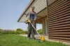 A woman is using a yellow and black string trimmer in a grassy yard next to a modern house with a brown slatted wall and a slatted roof overhang. Produktbild-Vorschau