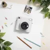A white Fujifilm instax SQUARE SQ 1 instant camera is placed on a white surface, surrounded by office supplies and greenery. Produktbild-Vorschau