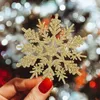 A hand with red nail polish holding a gold glitter snowflake ornament in front of a blurred background of Christmas lights. Náhľad obrázku produktu