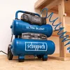 A blue Scheppach air compressor is shown in a workshop setting, with a wooden workbench in the background and a coiled blue air hose attached. Anteprima dell’immagine prodotto