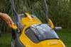 A person's hand is shown operating a yellow and gray lawnmower in an outdoor setting with green grass and tall plants in the background. Produktbild-Vorschau