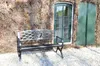 A black metal garden bench sits in front of a brick wall with ivy, next to a green door. Anteprima dell’immagine prodotto