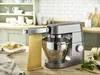 A Kenwood stand mixer with a pasta attachment is shown on a wooden countertop, with a baking dish and ingredients in the background. Produktbild-Vorschau