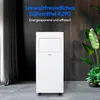 A white portable air conditioner is placed on a gray tiled floor in a room with dark wood paneling on the wall and a sheer curtain on the left. A plant is visible on the right. Produktbild-Vorschau