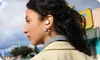 A woman wearing earbuds and a jacket, with a blurred background of buildings and trees. Produktbild-Vorschau