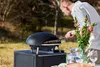 A man is using a black pizza oven outdoors on a table, with flowers and glasses nearby. Produktbild-Vorschau