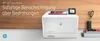 A white HP Color LaserJet Pro printer is shown on a desk with a wooden background. There are also some books, scissors, and a small screen on top of the printer. Podgląd zdjęcia produktu