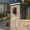 A black mailbox is mounted on a stone pillar next to a black metal fence, with a house in the background. Voir les images du produit