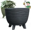 A dark grey planter with a textured surface and three legs is shown in the foreground, with a green planter containing a plant in the background. A cartoon illustration of a gardener giving a thumbs-up is in the upper right corner. Podgląd zdjęcia produktu