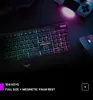 A gaming keyboard and mouse with RGB lighting on a dark surface, with gaming controllers in the background. Produktbild-Vorschau