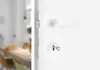 Close-up of a white door handle and lock mechanism on a white door, with a blurred interior scene visible through the doorway. Produktbild-Vorschau