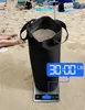 A black sandbag filled with sand sits on a scale on a beach, with beach chairs and umbrellas in the background. Produktbild-Vorschau