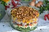 A layered salad in a glass bowl, with ingredients like broccoli, eggs, tomatoes, ham, corn, and peanuts visible. The salad is placed on a white wooden surface, with some decorative elements and eggs in the background. Podgląd zdjęcia produktu
