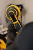 A person wearing gloves holds a yellow and black power tool, likely a sander, in a workshop or construction environment. Produktbild-Vorschau