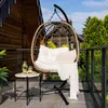 A hanging egg chair with cushions and a throw blanket is set up on a balcony, with a small side table holding a drink and a plate of food. Produktbild-Vorschau