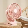A pink clip-on fan is placed on a wooden table. A wooden chair is visible in the background. Produktbild-Vorschau