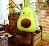 A plush avocado toy sits on a wooden crate with plants behind it. Produktbild-Vorschau