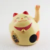 A light beige Maneki-neko (lucky cat) figurine with red ears and a red collar with white polka dots. It has a gold bell on its collar and black and red accents on its body. There is text on its left side. Produktbild-Vorschau