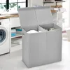 A laundry basket with two compartments and a lid, placed in a laundry room next to a washing machine and shelves with folded towels. Produktbild-Vorschau