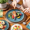 A person is eating a meal from a blue plate on a wooden table, with other dishes and a plant in the background. Náhled obrázku produktu