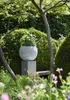 A garden scene with a planter on a pedestal, surrounded by greenery and trees. Náhľad obrázku produktu
