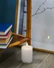 A white LED candle sits on a wooden floor next to a stack of books and a rattan shelf. Produktbild-Vorschau