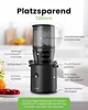 A black Hurom slow juicer is shown with labels pointing to its various parts and features, set against a blurred kitchen background. A small plate of strawberries and an orange are visible in the foreground. Podgląd zdjęcia produktu