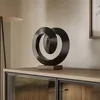 A decorative sculpture on a wooden cabinet, with a lamp and framed art in the background. Produktbild-Vorschau