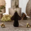 A small, dark brown paper mushroom decoration sits on a wooden surface, surrounded by natural elements like pinecones and dried flowers. Produktbild-Vorschau
